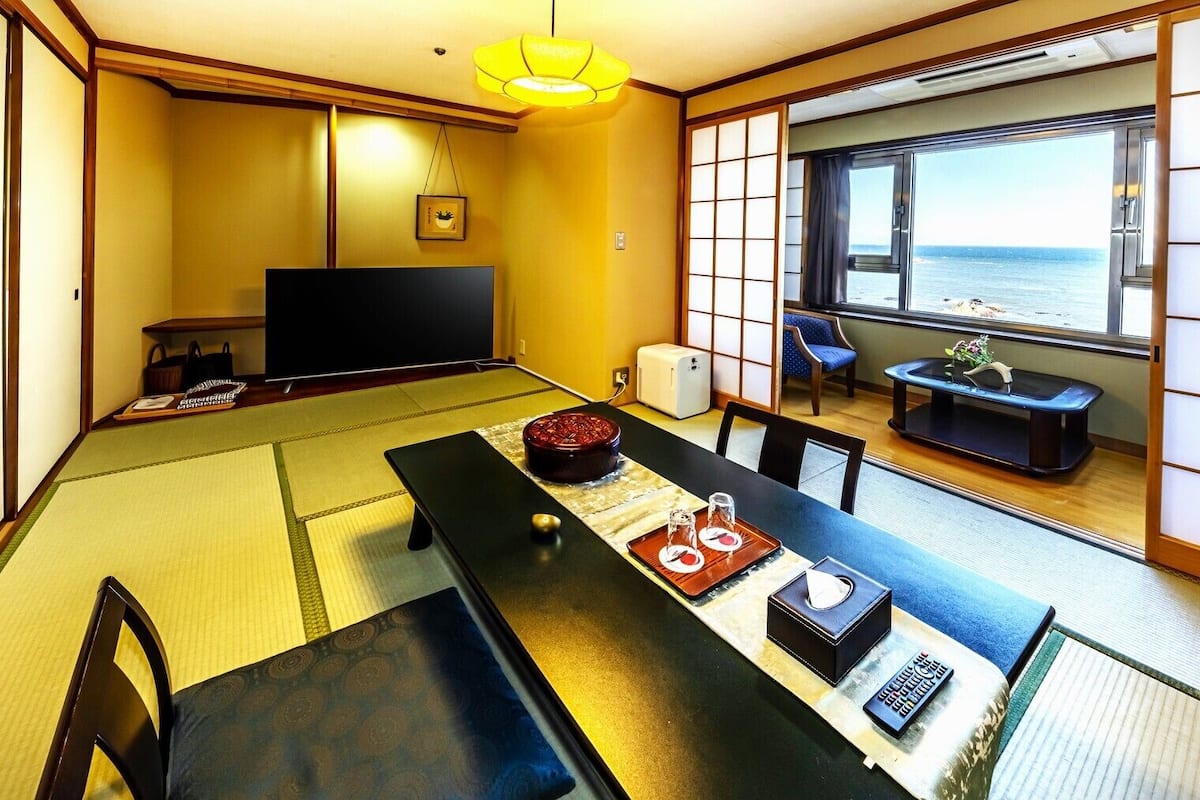 A traditional Japanese room with tatami flooring, a low table with a tea set, a television, and a view of the sea through a sliding door.