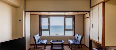 Ein Zimmer mit Tatami-Boden, einem Fernseher, zwei Ledersesseln, einem kleinen Tisch und Meerblick durch große Fenster.