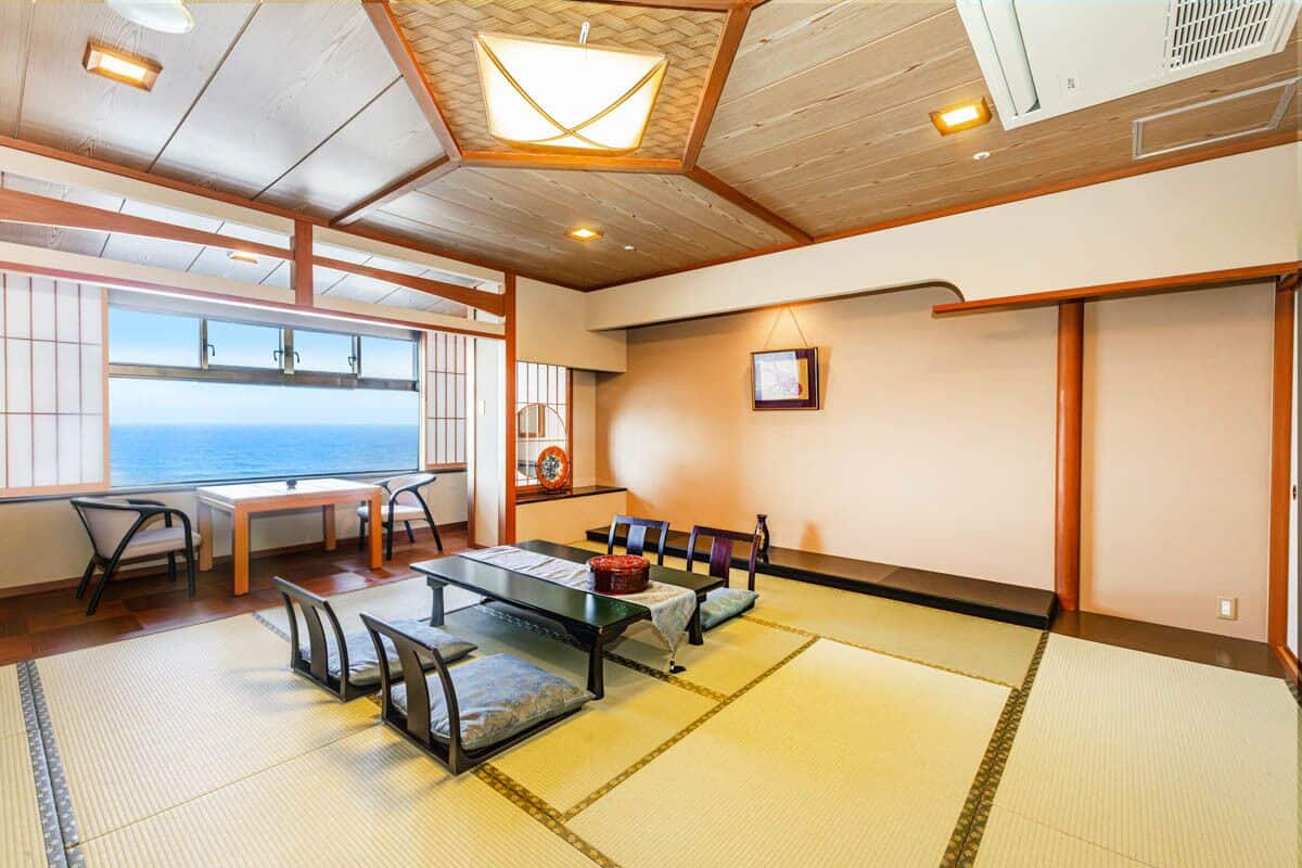 A traditional Japanese room with tatami flooring, a low table with cushions, and a view of the sea through large windows.