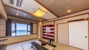 Une pièce de style japonais traditionnel, avec un sol recouvert de tatamis, une petite table avec un coussin, une vue sur la mer à travers de grandes fenêtres et des portes coulissantes ornées de motifs rouges et blancs.