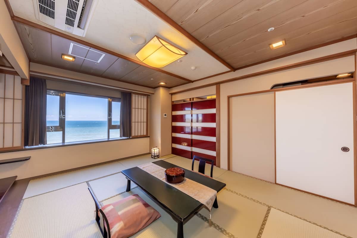 A traditional Japanese room with tatami flooring, a low table with a cushion, a view of the sea through large windows, and sliding doors with red and white patterns.
