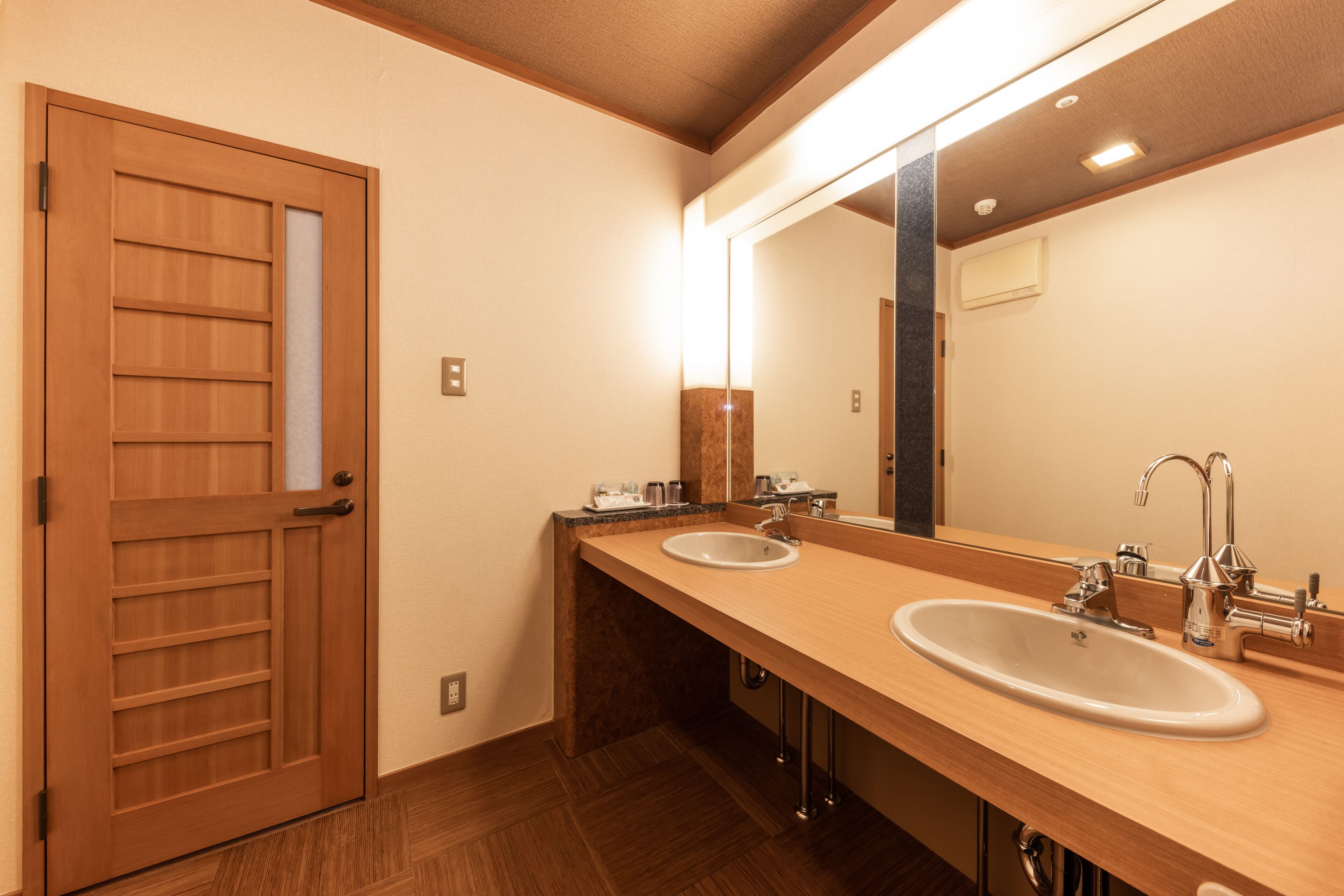 Un bagno d'albergo con pavimento in legno, due lavabi, uno specchio grande e una porta in legno.