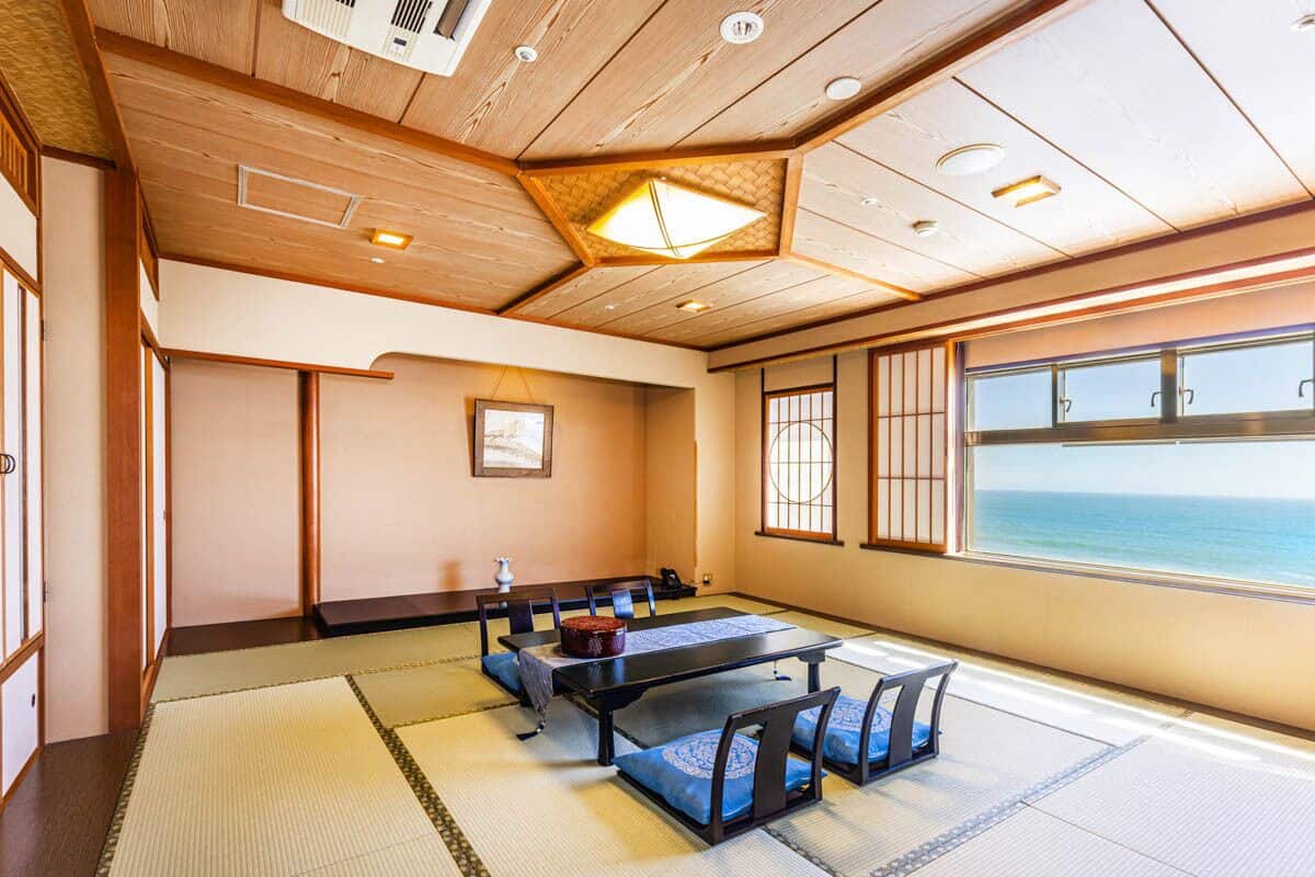 A traditional Japanese room with tatami flooring, a low table with cushions, and a view of the sea through large windows.