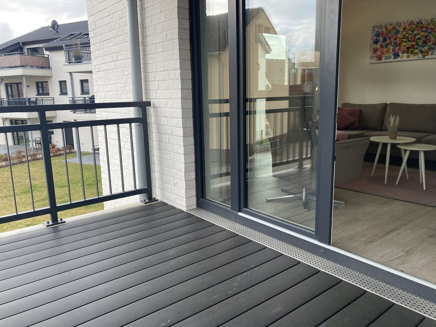Un balcon doté d’un plancher en bois, une porte vitrée donnant sur un espace de vie avec un canapé et une petite table, et une vue sur les maisons voisines.