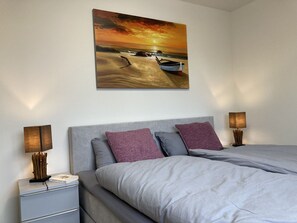 A bedroom with a bed, two bedside lamps, a nightstand, and a painting of a sunset over the ocean. - Dahmer Dünen - Küstenkönigin - Dahmer Dünen "küstenkönigin " - Strand- und Zentrumsnah! (Dahme)