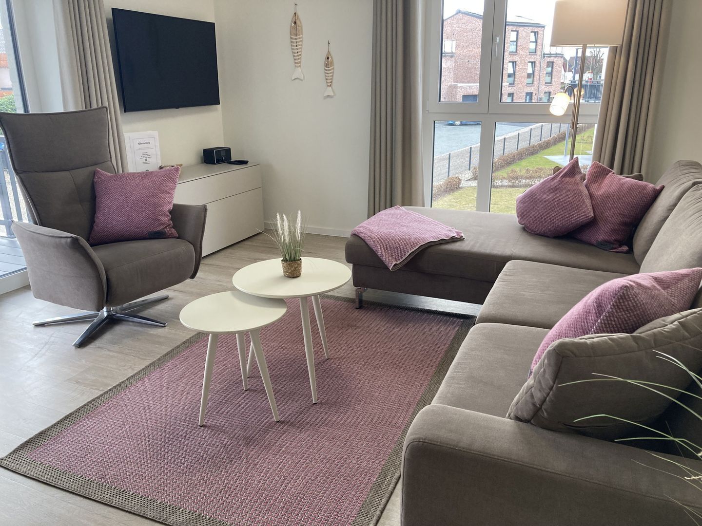 Een moderne woonkamer met een grijze bank, een bruine fauteuil en een witte salontafel. Op de salontafel staat een kleine plant in pot, en de kamer heeft grote ramen met uitzicht op gebouwen en een brug.