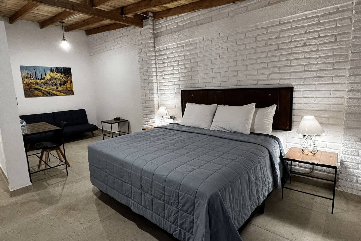 A bedroom with a large bed, a wooden headboard, a white brick wall, a small table with a lamp, a sofa, and a painting on the wall.