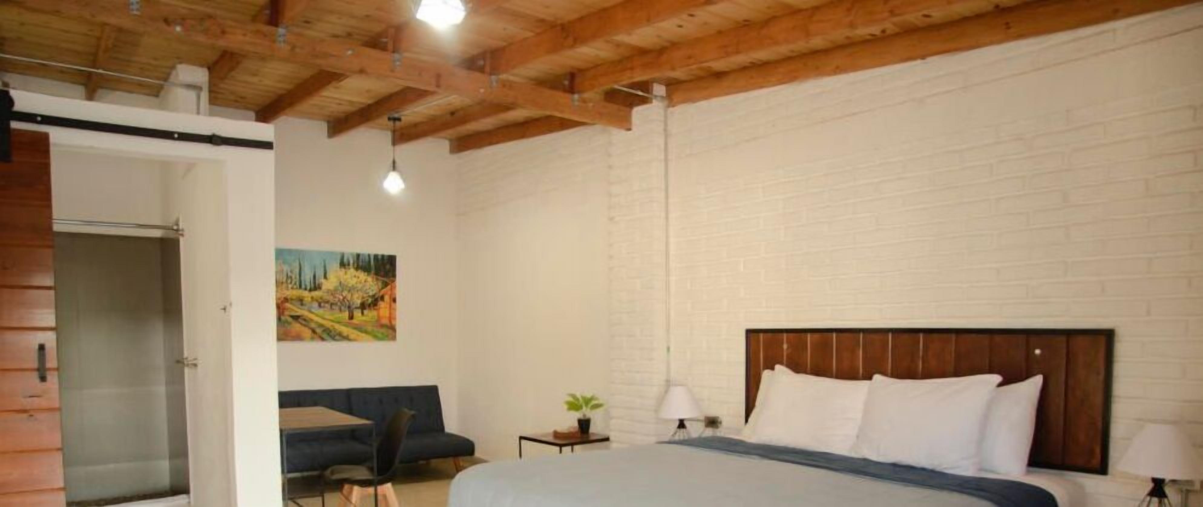 Un dormitorio con techo de madera, una cama con ropa de cama blanca, un sofá azul, una mesita de noche con una lámpara y un cuadro en la pared.