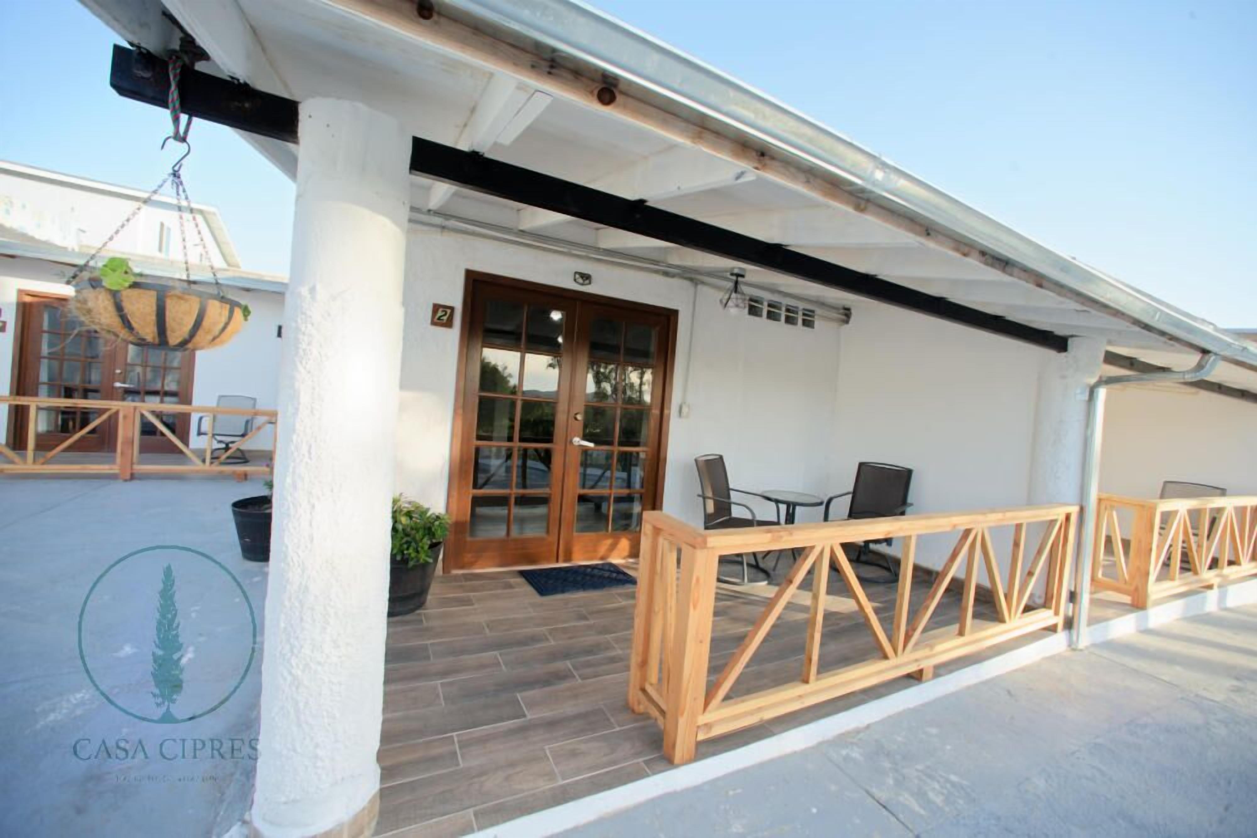 A covered outdoor area with a wooden table and chairs, a hanging basket, and a door leading to an interior space.