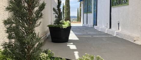 A narrow outdoor corridor with potted plants, a metal roof structure, and a view of a distant landscape.