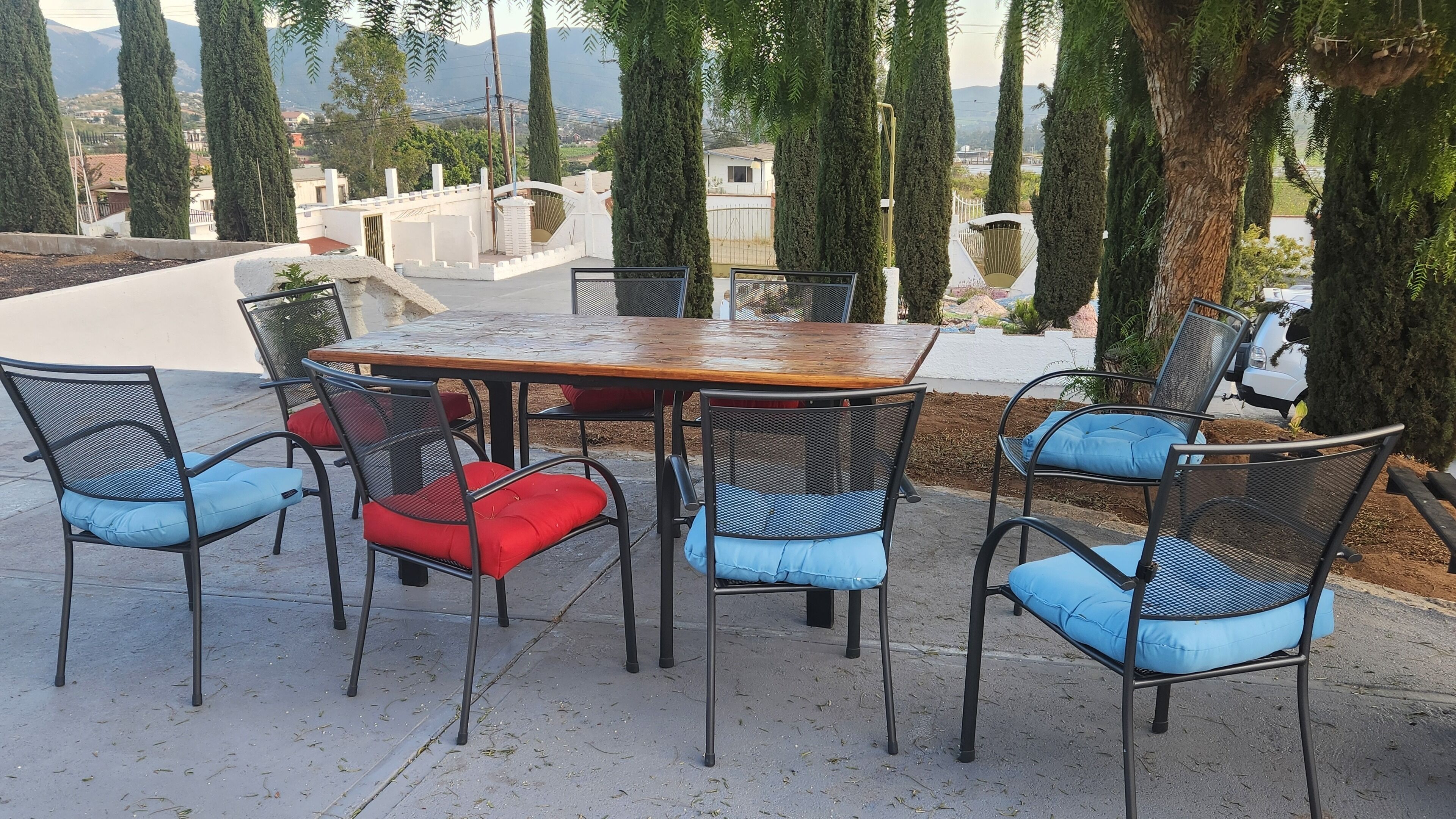 An outdoor seating area with a wooden table and chairs, surrounded by tall trees and a view of distant buildings.
