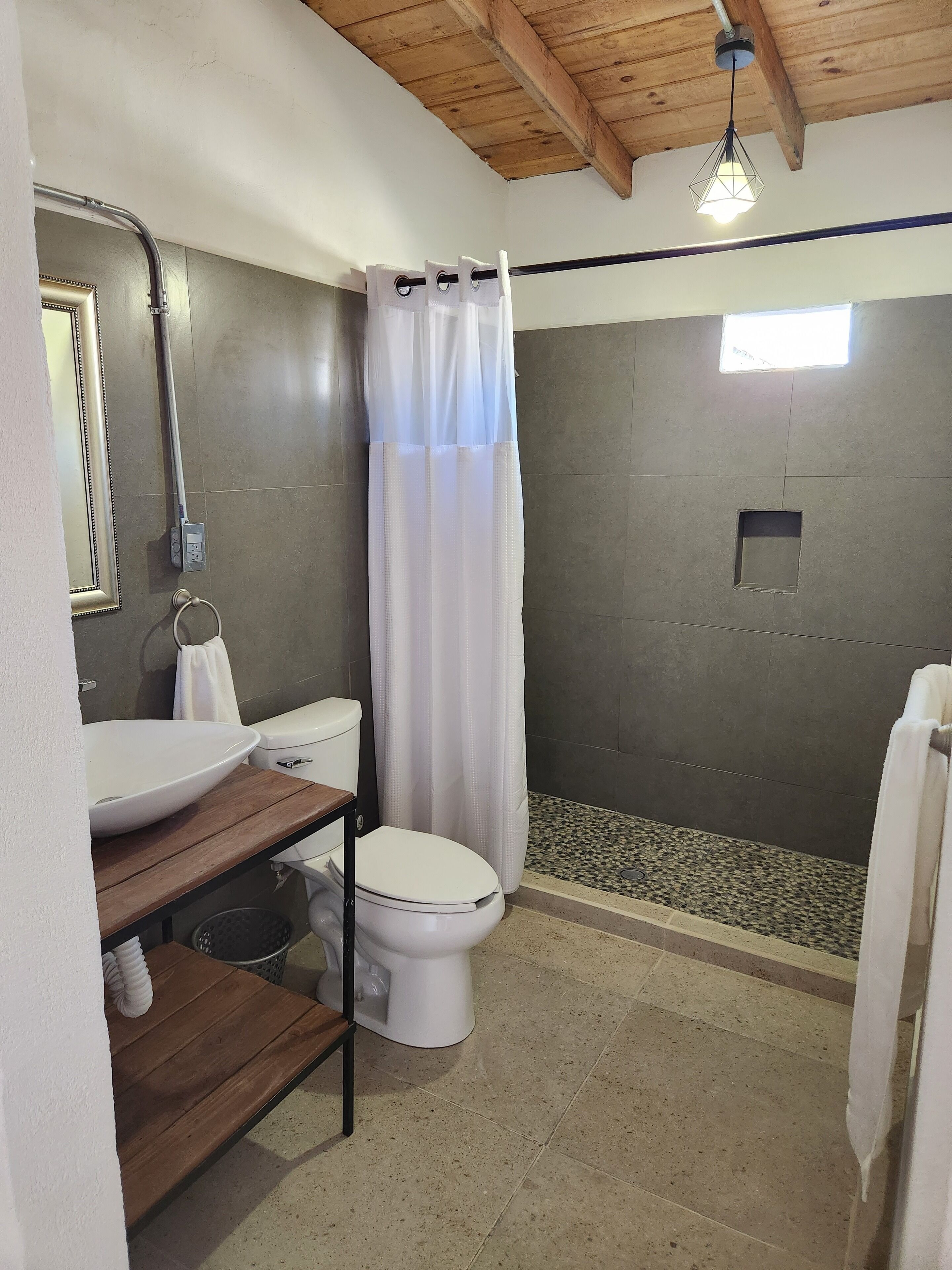 Une salle de bain avec un meuble-lavabo en bois, un lavabo blanc, des toilettes et un espace douche séparé par un rideau.