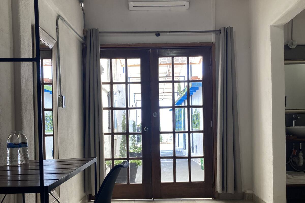 A room with a table, chair, and a large window with curtains. There is a door leading outside.