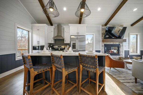 A modern kitchen with white cabinets, a large island with bar stools, a built-in oven, and a stone fireplace. - Couples, Hot Tub, Fire Pit, Pet Friendly, Peace & Quiet (Broken Bow)
