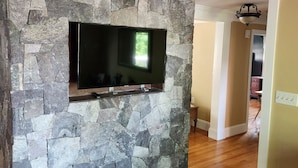 Un mur en pierre sur lequel est fixé un téléviseur à écran plat, un plancher en bois et un couloir menant à une autre pièce.