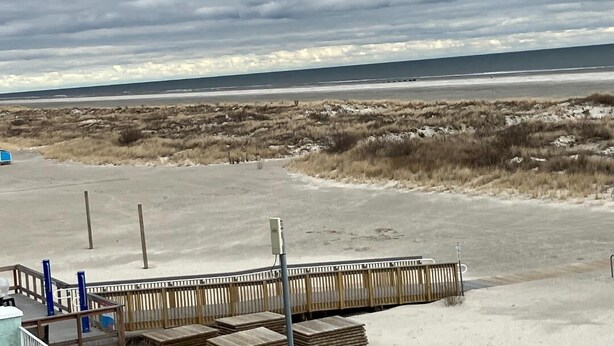 Una playa con un paseo marítimo de madera que lleva al agua, dunas de arena y un cielo nublado.