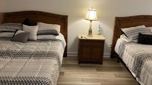 Habitación de hotel con dos camas, cada una con cabecero de madera, mesita de noche con lámpara y un mueble auxiliar.