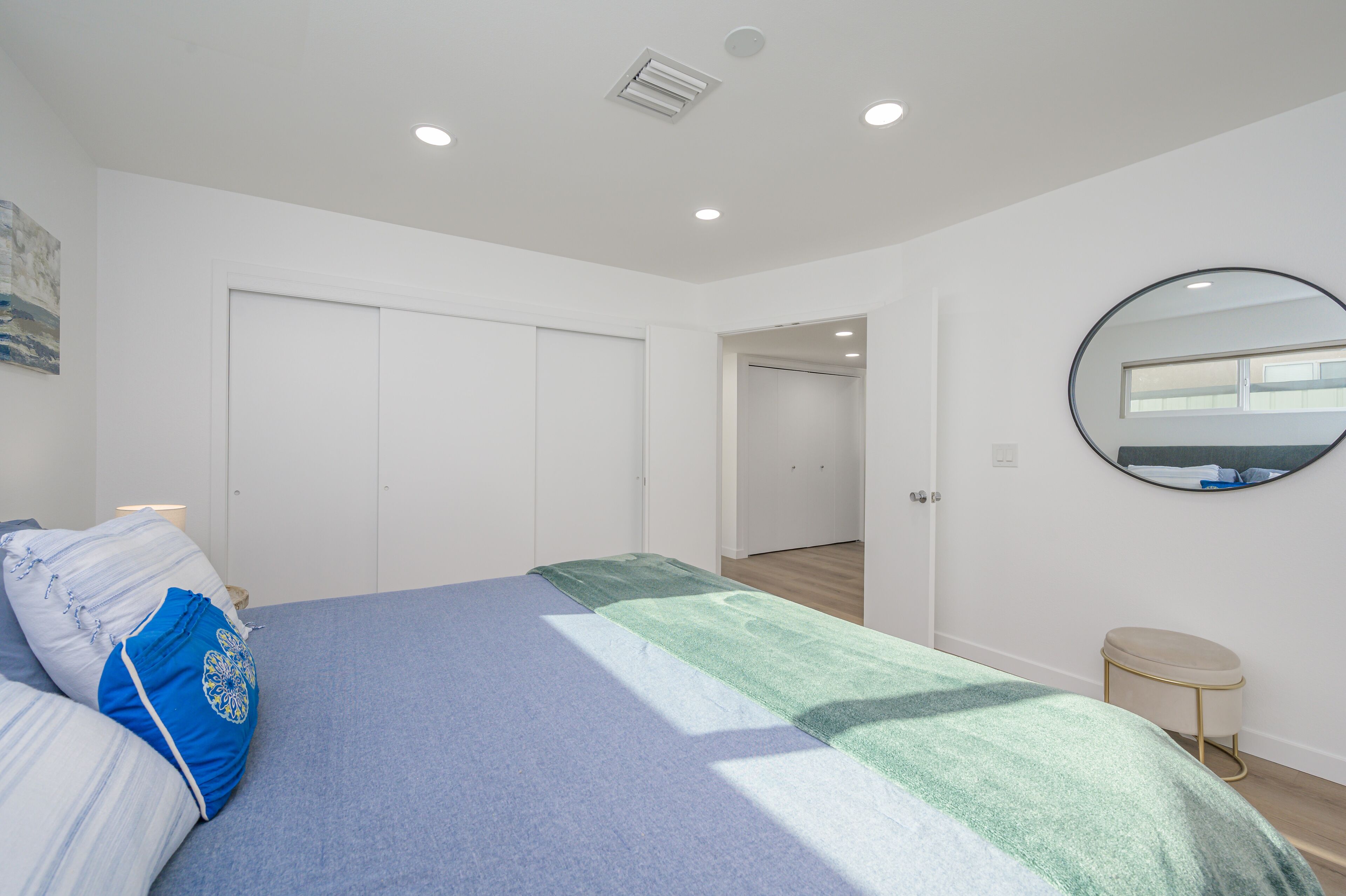 Une chambre à coucher avec un lit, un miroir rond, un petit tabouret et une porte donnant sur une autre pièce.