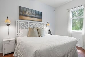 A bedroom with a large bed, two bedside lamps, a white nightstand, a decorative headboard, and a window with a view of greenery.
