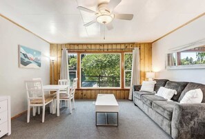 Une pièce comprenant une table à manger, des chaises, un canapé, un ventilateur de plafond et de grandes fenêtres donnant sur un paysage verdoyant.