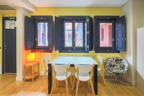 Un comedor con una mesa y sillas, dos ventanas con contraventanas, una puerta con la etiqueta 'SALIDA' y una lámpara pequeña sobre una mesita.
