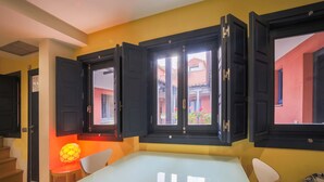 Un comedor con una mesa y sillas, una ventana con contraventanas abiertas y vistas a un edificio rojo.