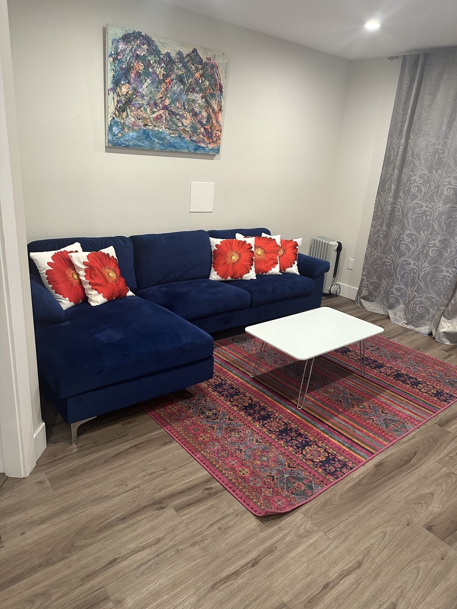 A modern living room with a blue sofa, red floral cushions, a white coffee table, a patterned rug, and a large abstract painting on the wall.