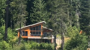A modern house with a large window, surrounded by dense forest and a body of water.
