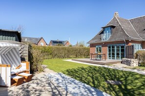 Una propiedad residencial con césped cuidado, un patio con muebles de exterior y una casa de ladrillo con ventanas amplias y techo inclinado.