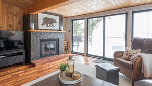 A cozy living room with a stone fireplace, wooden ceiling, and large windows. - Waterfront Cottage on the Lake with Game Room & Sunroom (Lakefield)