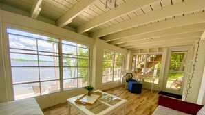 A spacious room with large windows overlooking a body of water, a wooden floor, and a white ceiling. - Waterfront Cottage on the Lake with Game Room & Sunroom (Lakefield)