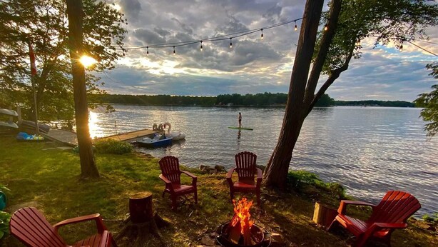 A lakeside outdoor seating area with Adirondack chairs, a fire pit, and a dock with a boat. - Waterfront Cottage on the Lake with Game Room & Sunroom (Lakefield)
