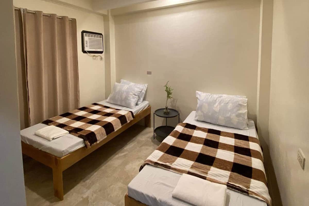 A small, clean room with two single beds, a small table, and a ceiling light.
