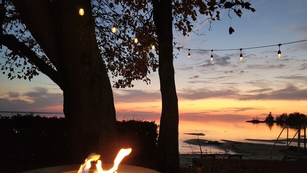 A fire pit with flames, surrounded by trees and a string of lights against a sunset backdrop. - Cozy Beachfront Cottage (North Bay)