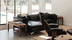 Un salon spacieux avec un canapé en cuir noir, une table basse en bois et un ventilateur de plafond. De grandes fenêtres laissent entrer la lumière naturelle et offrent une vue sur l’extérieur.