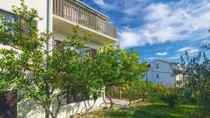 A two-story residential building with a balcony, surrounded by lush greenery and fruit trees.