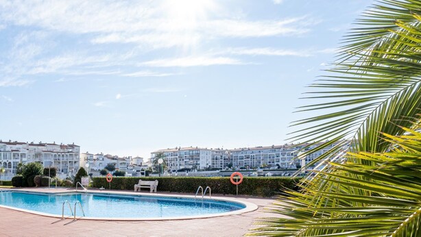 A swimming pool surrounded by a well-maintained lawn and a row of white apartment buildings. - Lake view apartment with 1 bedroom and community pool (Empuriabrava)