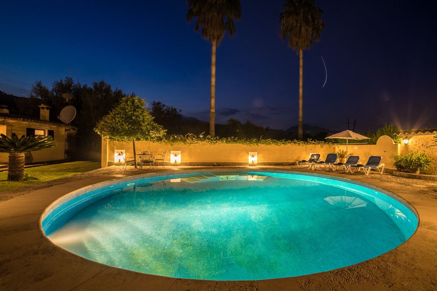 A swimming pool with lounge chairs and a table, surrounded by palm trees and a building with satellite dishes.