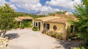 A traditional Mediterranean-style villa with a paved driveway leading to a gated entrance, surrounded by lush greenery and a mountainous backdrop. - 3 bedroom country house with private pool and barbecue (Pollensa)