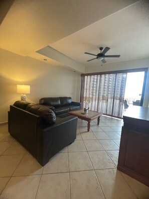 A living room with a black leather sofa, a wooden coffee table, a ceiling fan, and a large window with a view of the outside.