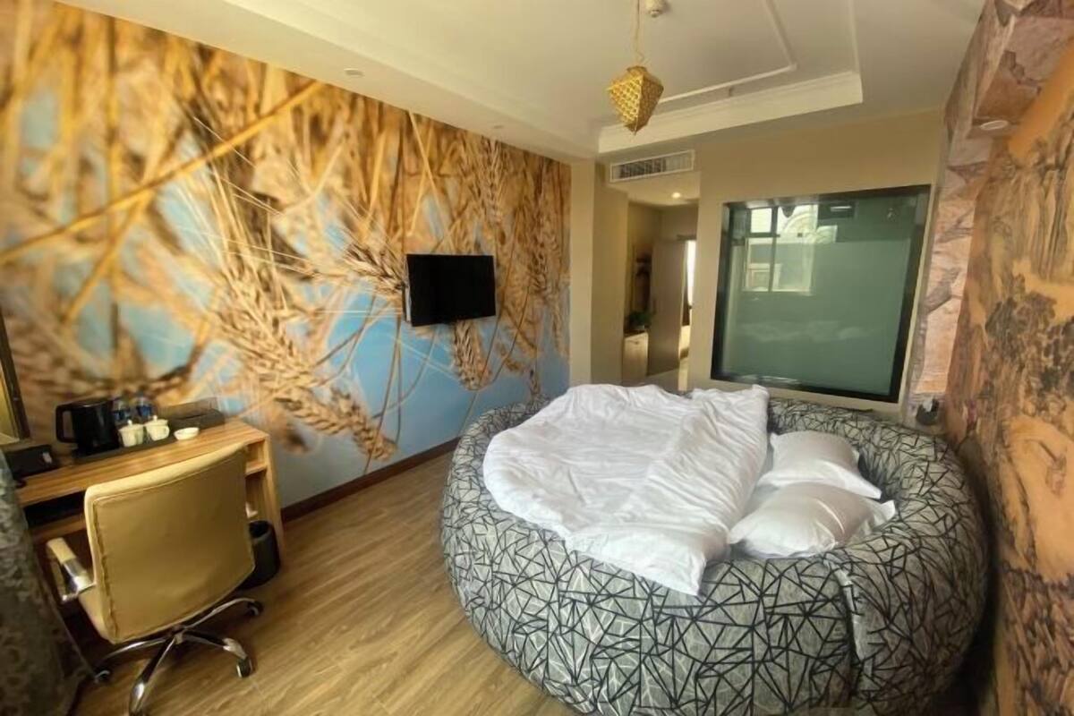 Une chambre d’hôtel avec un lit rond, un bureau avec une chaise, une télévision et une grande fenêtre.