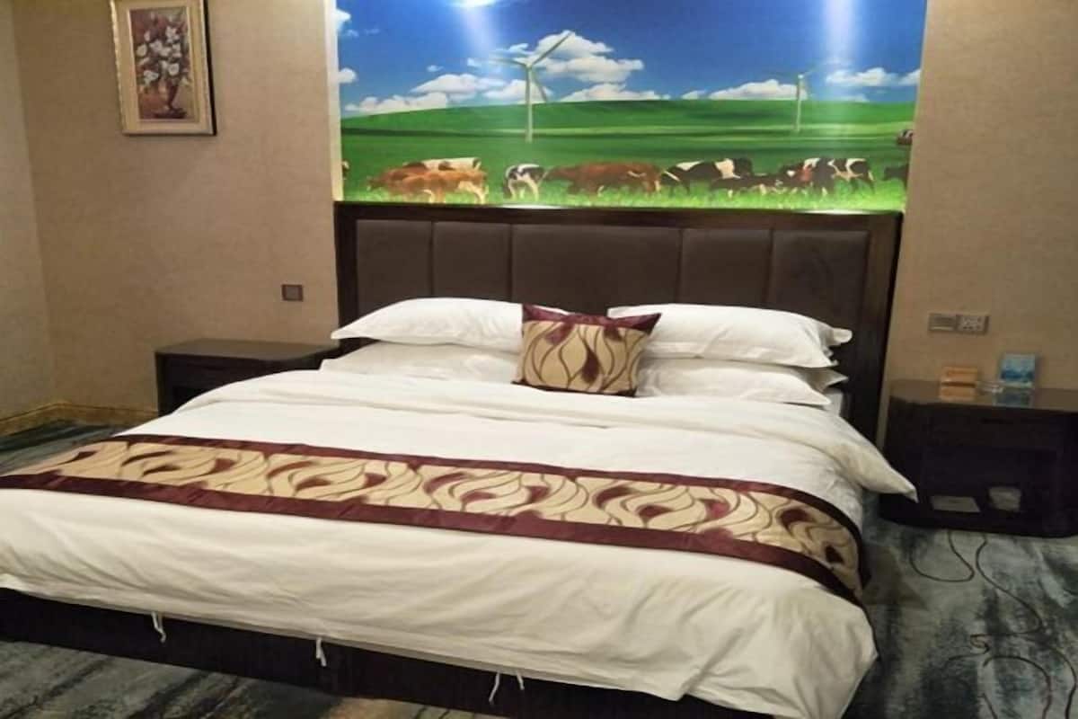 Une chambre d’hôtel avec un lit, une table de chevet, un téléviseur fixé au mur qui diffuse une scène champêtre, et un tableau encadré accroché au mur.