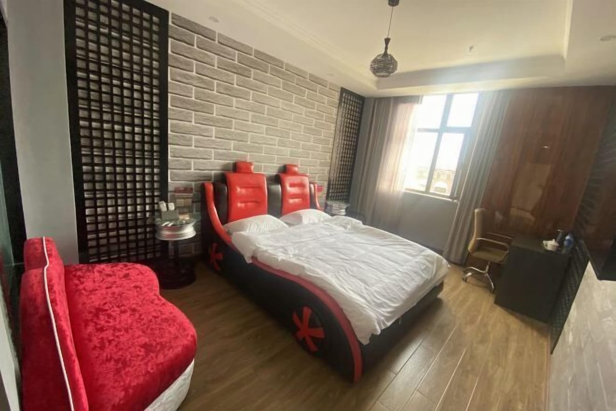 A room with a bed, a red chair, a small table, and a window.