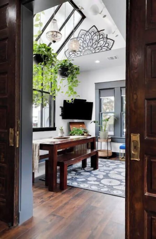 A room with a wooden floor, a dining table with chairs, a bench, hanging plants, a television mounted on the wall, and a patterned rug.