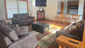 A living room with a sofa, a coffee table, and a TV. There is a kitchen area with a microwave and a dining table with chairs.