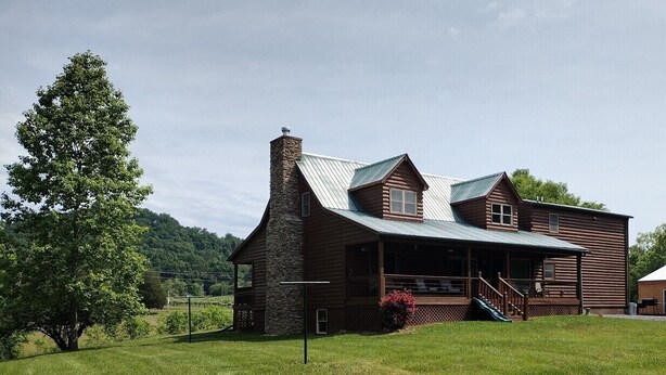 A rustic log cabin with a stone chimney, a porch, and a large tree in the foreground.