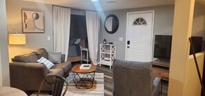 Una sala de estar con sofá, sillón, mesa de centro y televisor. Se ve una puerta, un espejo y una lámpara.