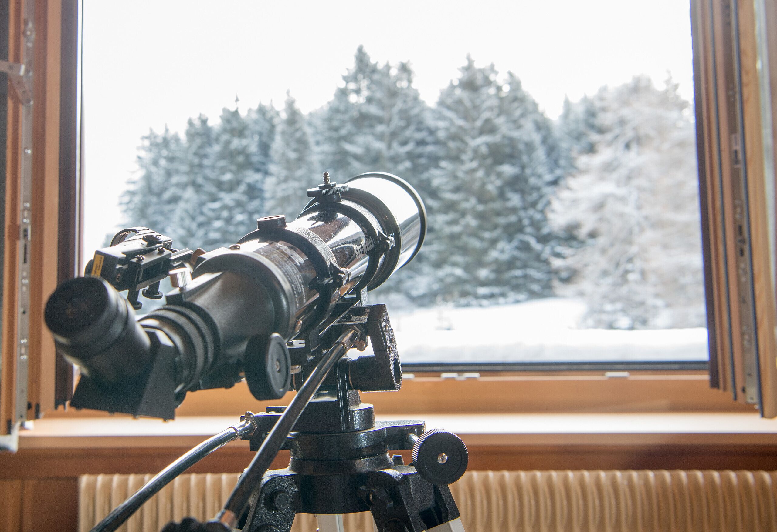 Un telescopio sobre un trípode junto a una ventana con vista a un bosque nevado.