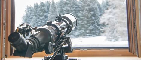Un telescopio sobre un trípode junto a una ventana con vista a un bosque nevado.