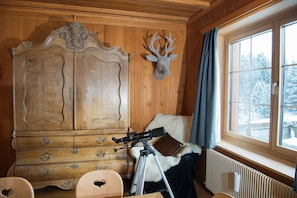 A wooden room with a large cabinet, a mounted deer head, a telescope on a tripod, a fur-covered chair, and a window with a view of snow-covered trees.